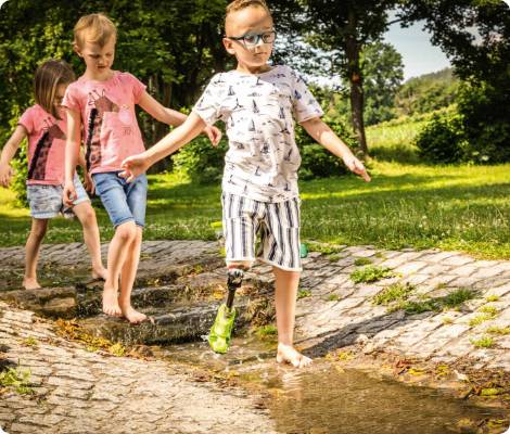 Auf dem Bild sind drei Kinder zu sehen, die durch einen Wasserlauf laufen und ein Kind trägt eine Unterschenkelprothese.